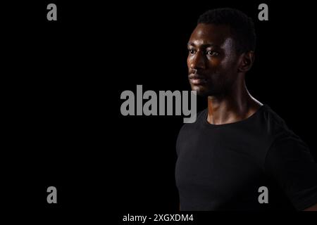 Homme afro-américain dans une chemise noire pose soigneusement sur un fond noir. Son expression sérieuse transmet un sentiment de détermination et de concentration. Banque D'Images