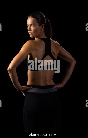 Jeune athlète féminine caucasienne pose avec confiance en vêtements de sport sur un fond noir. Sa forme athlétique est mise en évidence sur un fond sombre, emph Banque D'Images