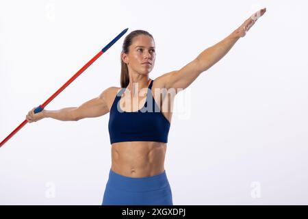 Une jeune athlète féminine caucasienne se prépare à lancer un javelot sur un fond blanc. Son expression ciblée et sa position athlétique soulignent l'intensité O. Banque D'Images