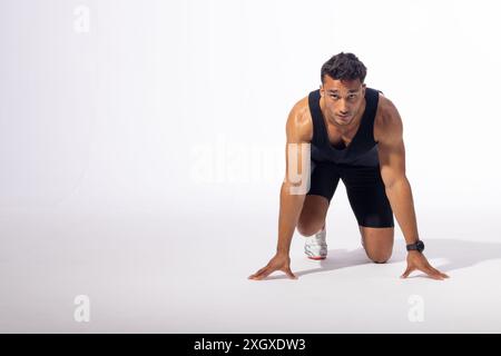 Jeune homme biracial dans la position de départ d'un coureur, avec espace de copie sur fond blanc. Son expression focalisée et sa pose athlétique suggèrent la déterminatio Banque D'Images