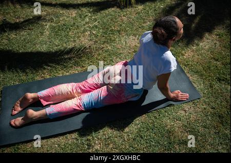 Femme s'engageant dans une séance de yoga en plein air sur une journée ensoleillée, effectuant la pose du chien face vers le haut. Elle est sur un tapis de yoga, portant des pantalons teints vibrants. Banque D'Images