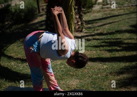 Femme pratiquant le yoga en plein air dans le parc, effectuant une pose penchée vers l'avant avec ses mains serrées sous le ciel ouvert et la lumière chaude du soleil. Banque D'Images