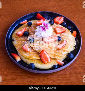 Délicieuses crêpes garnies de crème fouettée, de myrtilles et de fraises tranchées servies sur une assiette noire Banque D'Images