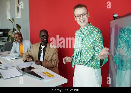 Équipe de professionnels divers pendant la réunion avec le présentateur faisant des gestes vers l'écran pendant que les collègues écoutent attentivement. Discussion de groupe engagée dans un environnement de bureau moderne Banque D'Images