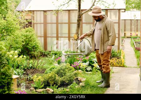 Vue latérale de l'agriculteur mâle mature avec pot d'arrosage rafraîchissant plantes vertes poussant sur un grand parterre de fleurs tout en se tenant sur un chemin en béton Banque D'Images