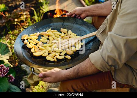 Plan recadré de jardinier masculin mature dans des vêtements de travail tenant une grande poêle à frire chaude ronde avec des pommes de terre crues hachées et une spatule en bois Banque D'Images