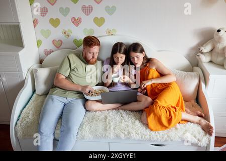 Famille de trois est assis sur un canapé blanc, regardant quelque chose sur un ordinateur portable tout en grignotant Banque D'Images