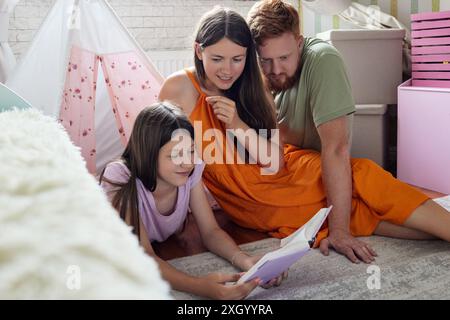 La jeune fille lit un livre avec ses parents dans une salle de jeux, entourée de jouets et d'une tente confortable Banque D'Images