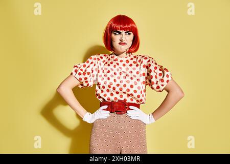 Femme aux cheveux rouges vibrants avec un maquillage pop art enfilant une blouse à pois sur un fond jaune audacieux. Banque D'Images