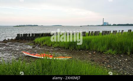 NORWALK, CT, USA - 9 JUILLET 2024 : un kayak orange vif repose sur la rive rocheuse près d'une zone herbeuse le soir d'été Banque D'Images