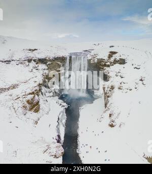 Vue aérienne de la cascade de Skogafoss en Islande en hiver, entourée de paysages enneigés avec des visiteurs à la base. Banque D'Images