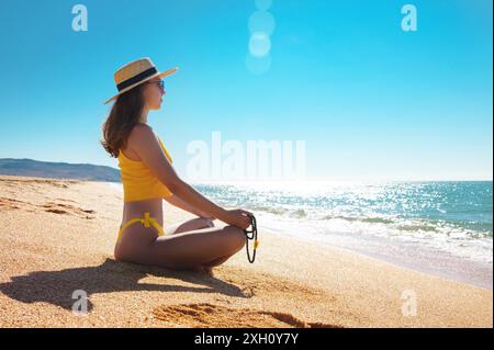 Méditation au bord de la mer par une journée ensoleillée. Jeune femme caucasienne dans un maillot de bain jaune est assise sur le sable doré dans une position de lotus dans un chapeau de paille et Banque D'Images