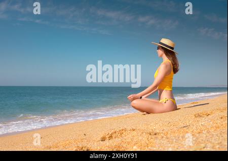 Méditation au bord de la mer par une journée ensoleillée. Jeune femme caucasienne dans un maillot de bain jaune est assise sur le sable doré dans une position de lotus dans un chapeau de paille et Banque D'Images