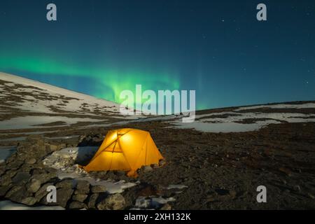 Ciel étoilé de nuit et aurores boréales vertes au-dessus de la tente jaune incandescente dans les montagnes Khibiny la nuit d'hiver. Russie. Banque D'Images