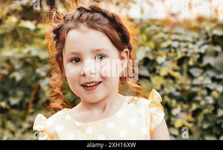 Jolie petite fille avec des cheveux bouclés souriant et regardant caméra en vous tenant contre les buissons le jour de l'été dans le jardin Banque D'Images