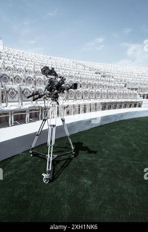 Une caméra vidéo professionnelle sur trépied est installée devant des rangées de chaises vides ornées dans un lieu extérieur sous un ciel bleu clair. La scène Suge Banque D'Images