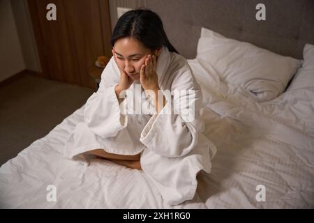 Femme asiatique bouleversée assise sur le lit dans un peignoir se penchant la tête sur les mains Banque D'Images