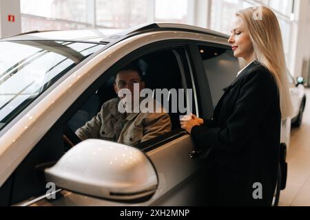 Vue arrière du concessionnaire automobile féminin en costume d'affaires parlant au jeune client homme assis dans le salon de voiture. Gestionnaire professionnel aidant le client masculin à choisir Banque D'Images