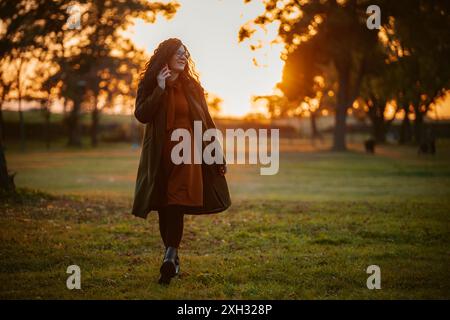 Une femme en long manteau marche à travers un parc herbeux au coucher du soleil. La lumière dorée illumine la scène alors qu'elle marche vers l'horizon. Banque D'Images