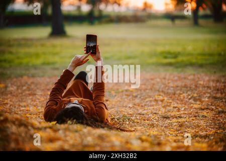 Une femme est allongée sur le dos dans un parc, entourée de feuilles tombées. Elle utilise son smartphone. Banque D'Images