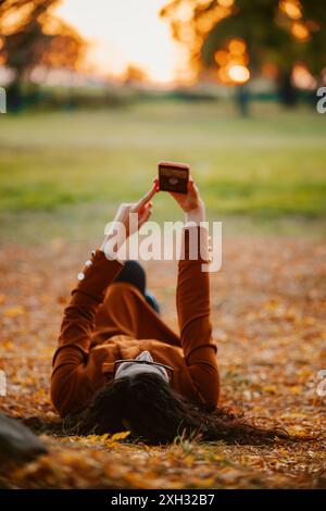 Une femme portant un manteau brun est couchée sur le dos dans un parc, entourée de feuilles tombées, en utilisant son smartphone. Banque D'Images