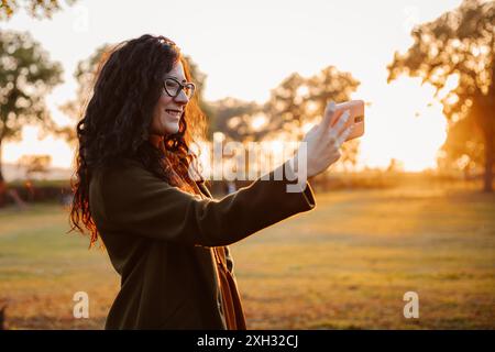 Une femme aux longs cheveux bouclés prend un selfie avec son smartphone tout en se tenant dans un parc au coucher du soleil. Le soleil brille à travers les arbres en arrière-plan Banque D'Images