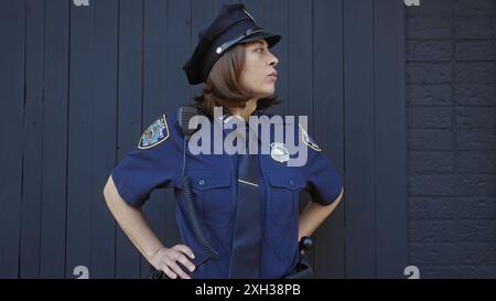 Une policière sérieuse en uniforme se tient contre un mur urbain sombre, transmettant autorité et sécurité. Banque D'Images
