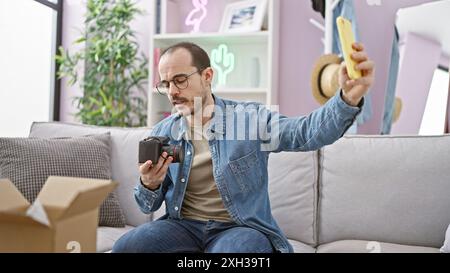 Homme hispanique avec barbe enregistrant une vidéo de déballage sur smartphone dans le salon. Banque D'Images
