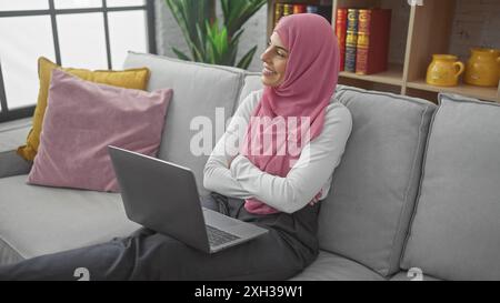 Une jeune femme souriante dans un hijab rose est assise les bras croisés sur un canapé gris à la maison, ordinateur portable à côté d'elle. Banque D'Images