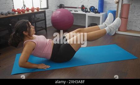 Une jeune femme hispanique attrayante exerçant à l'intérieur dans une salle de gym sur un tapis bleu effectuant des levées de jambes. Banque D'Images