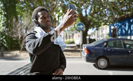 Jeune homme afro-américain adulte prend un selfie tout en portant des écouteurs à l'extérieur dans un cadre urbain ensoleillé. Banque D'Images