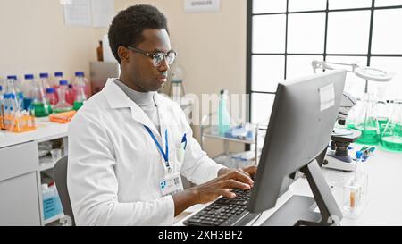 Un homme afro-américain concentré portant une blouse de laboratoire travaille sur un ordinateur dans un cadre de laboratoire lumineux. Banque D'Images