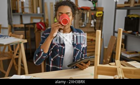 Jeune femme aux cheveux bouclés buvant du café dans un atelier de menuiserie tout en tenant une tablette. Banque D'Images