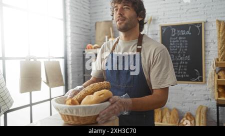 Jeune homme dans une boulangerie tenant un panier de pain, portant un tablier bleu et debout devant un tableau de menu avec des pains exposés Banque D'Images