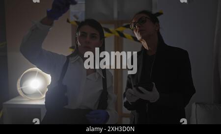 Deux enquêteurs femmes analysent les preuves dans une scène de crime intérieure faiblement éclairée. Banque D'Images