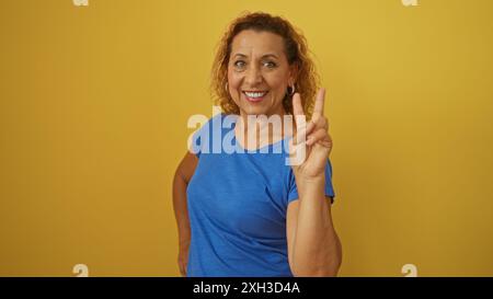 Belle femme hispanique âgée en chemise bleue, posant avec confiance avec un geste de signe de paix, isolée sur un fond jaune vif, incarnant Banque D'Images