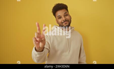 Bel homme noir faisant signe de paix en sweat à capuche beige sur fond jaune Banque D'Images