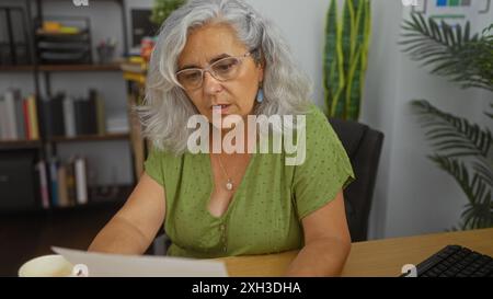 Femme âgée, aux cheveux gris, lisant des documents à un bureau, avec des étagères et des plantes en arrière-plan, concentrée sur le travail Banque D'Images