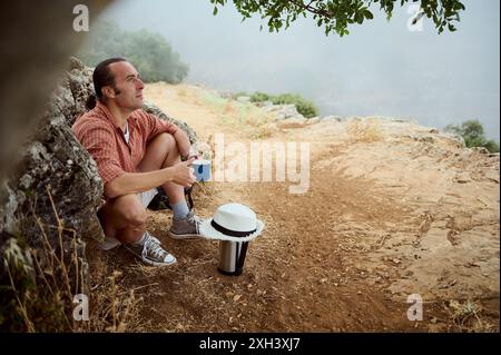 Un homme portant des vêtements décontractés est assis sur un chemin rocheux dans la nature, profitant d'un moment de tranquillité avec une tasse de café. La scène dépeint un sentiment de détente Banque D'Images