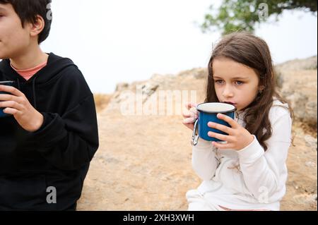 Deux enfants apprécient les boissons chaudes des tasses tout en étant assis sur un paysage rocheux en plein air. Un garçon dans un sweat à capuche noir et une fille dans une veste blanche. Banque D'Images