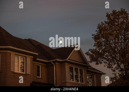 Maison de banlieue éclairée par le coucher du soleil - croissant de lune au-dessus - teintes chaudes sur la façade en brique - motifs complexes des fenêtres - arbre d'automne au flou doux. Prise à Toronto Banque D'Images