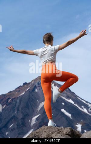 Vue arrière d'une femme pratiquant le yoga, debout sur le sommet d'une montagne rocheuse avec un volcan en arrière-plan. Femme galbée équilibre le corps sur une jambe. Professeur de yoga Banque D'Images