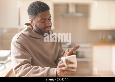 Homme heureux dans le sweat à capuche brun en utilisant le téléphone, étudier, apprendre, faire des achats, travailler en ligne, parler en étant assis sur un canapé à la maison Banque D'Images