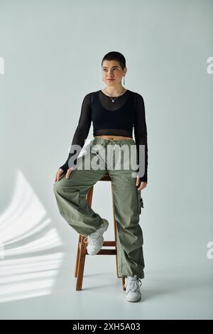 Vêtue d'une tenue chic, une jeune femme aux cheveux courts s'assoit gracieusement sur un tabouret en bois élégant sur un fond gris. Banque D'Images