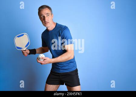 Portrait de joueur de pickleball prêt à faire un service avec une boule orange dans sa main sur fond isolé bleu. Banque D'Images