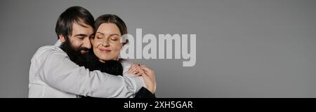 Un homme avec une barbe embrasse une femme, tous deux vêtus de vêtements sophistiqués, dans un moment tendre sur un fond gris. Banque D'Images