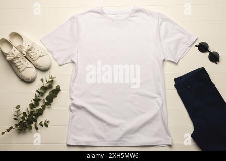 Un t-shirt blanc Uni est disposé à plat à côté de baskets blanches décontractées, lunettes de soleil foncées et pantalon bleu marine, avec une branche de feuilles vertes ajoutant une touc Banque D'Images