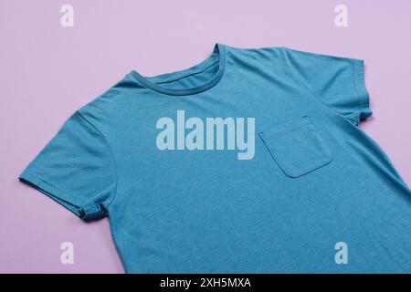 Un t-shirt bleu Uni avec une poche est disposé sur un fond violet, avec un espace de copie. Sa coupe simple en fait un vêtement décontracté ou customizat Banque D'Images