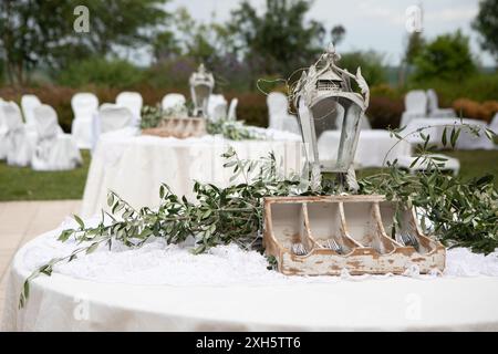 Table ronde décorée d'une nappe blanche, de branches d'olivier, d'argenterie et d'une lanterne à bougie vintage pour une réception de mariage en plein air Banque D'Images