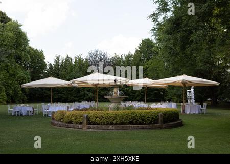 Préparation d'une réception de mariage avec des tables rondes élégantes et des chaises blanches dans un jardin avec une fontaine Banque D'Images